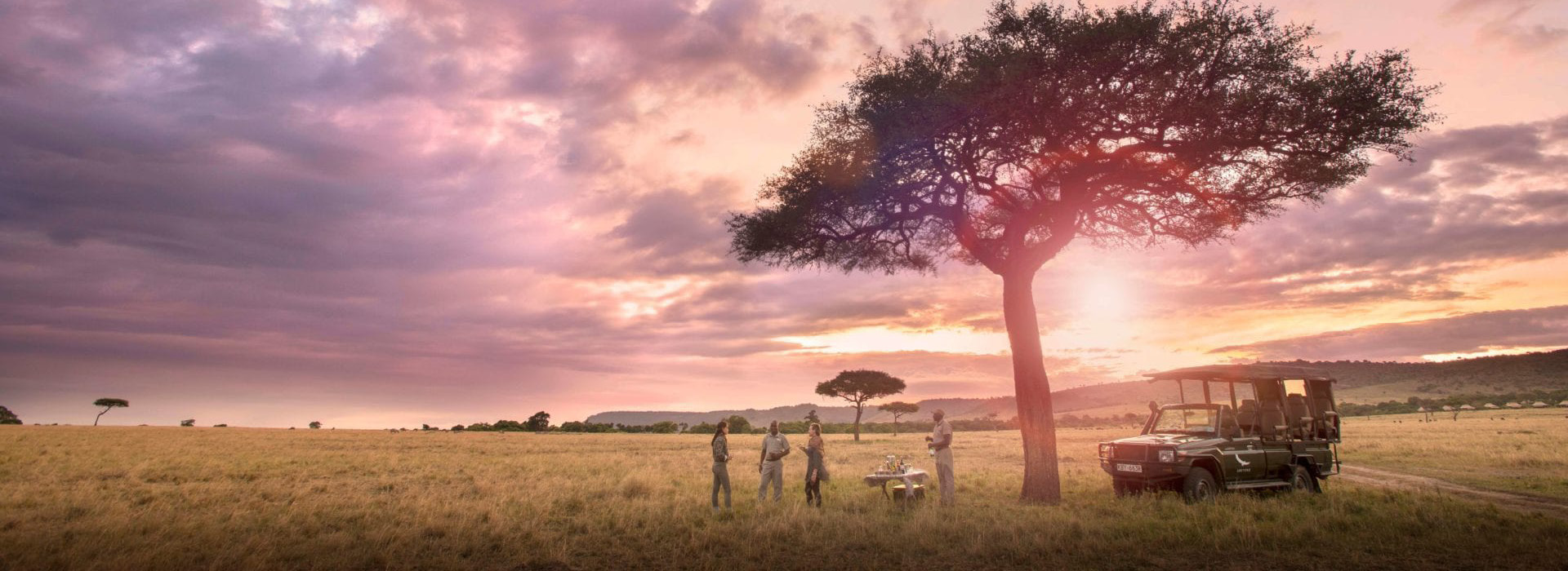 Safari vehicle and guests enjoying sundowners at a camp in the Maasai Mara, with a breathtaking sunset over the African savanna.